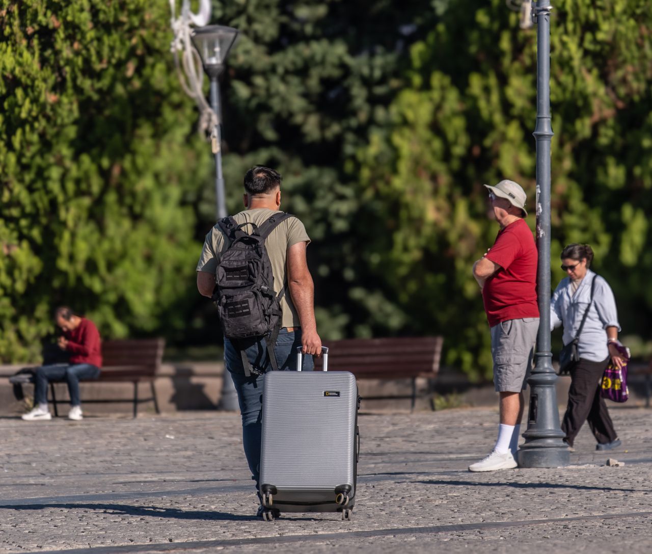 A man with a suitcase and backpack strolling in a sunny park in İzmir, Türkiye.